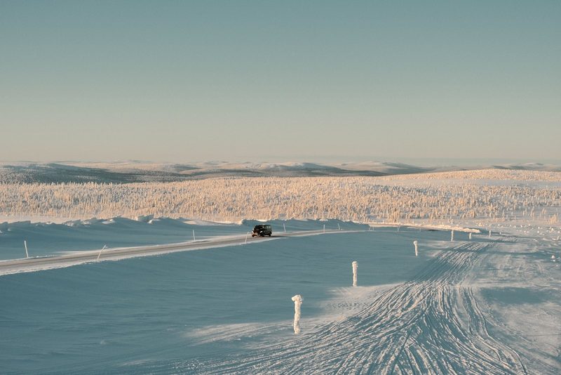A black SUV drives down a snow-covered road, flanked by a snow-covered field and a clear blue sky.