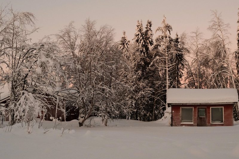 A small, red, one-story building with a peaked roof and three windows is situated in a snowy landscape, surrounded by trees and snow-covered ground.