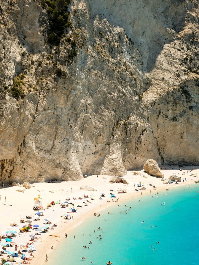 A large cliff rises behind a sandy beach, where people are gathered and swimming in the turquoise water.