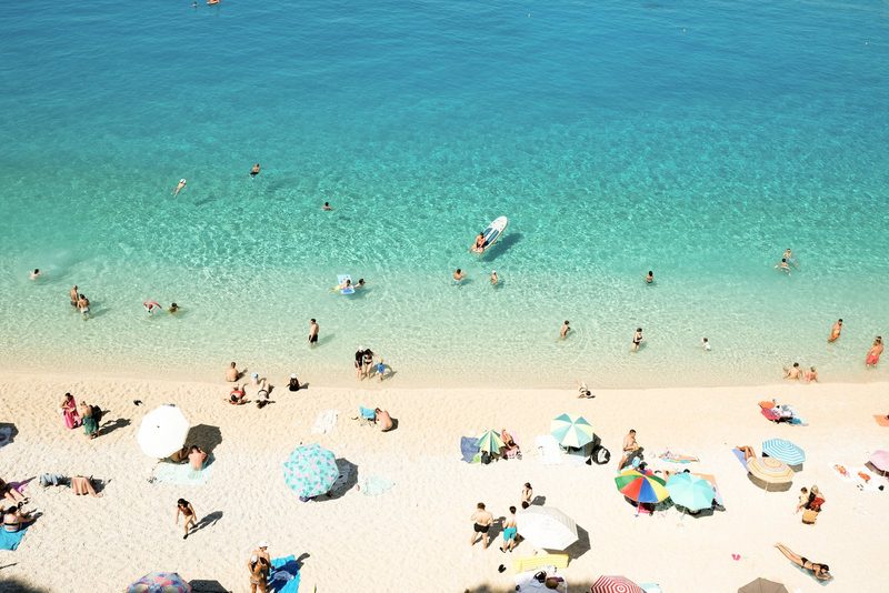 A beach scene with a large body of water, featuring people swimming and sunbathing on the shore, with various beach umbrellas and towels scattered about.