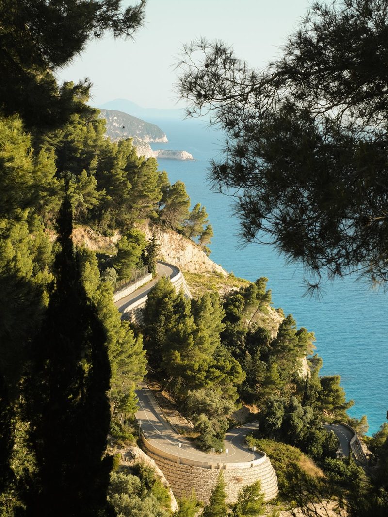 A winding road, lined with trees and a stone wall, runs along the edge of a cliff overlooking a body of water.