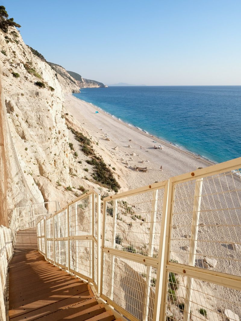 A long, narrow wooden walkway with a metal railing runs along the side of a steep cliff, overlooking a sandy beach and the ocean.