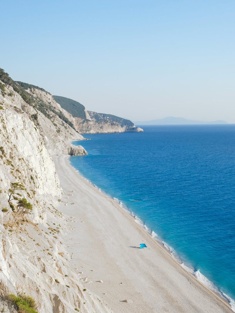 A sandy beach with a steep cliff on the left, a calm sea with a few waves, and a small blue object on the sand.