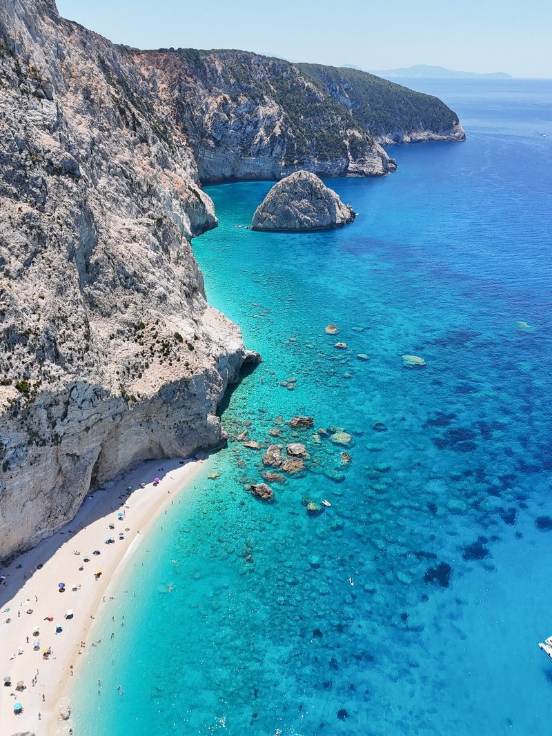 A rocky cliffside overlooks a sandy beach and a crystal-clear body of water, with a few boats and people scattered about.