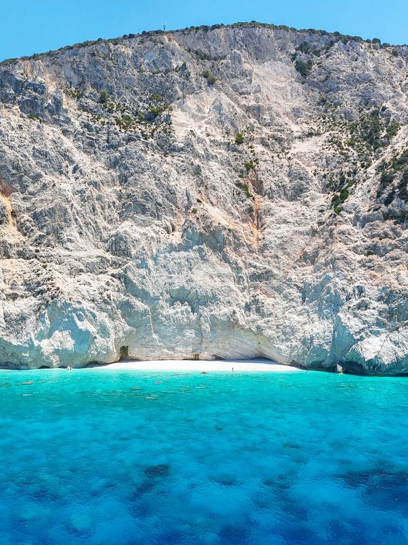 A large rocky cliff rises from a bright blue body of water, with a small white beach nestled at its base.