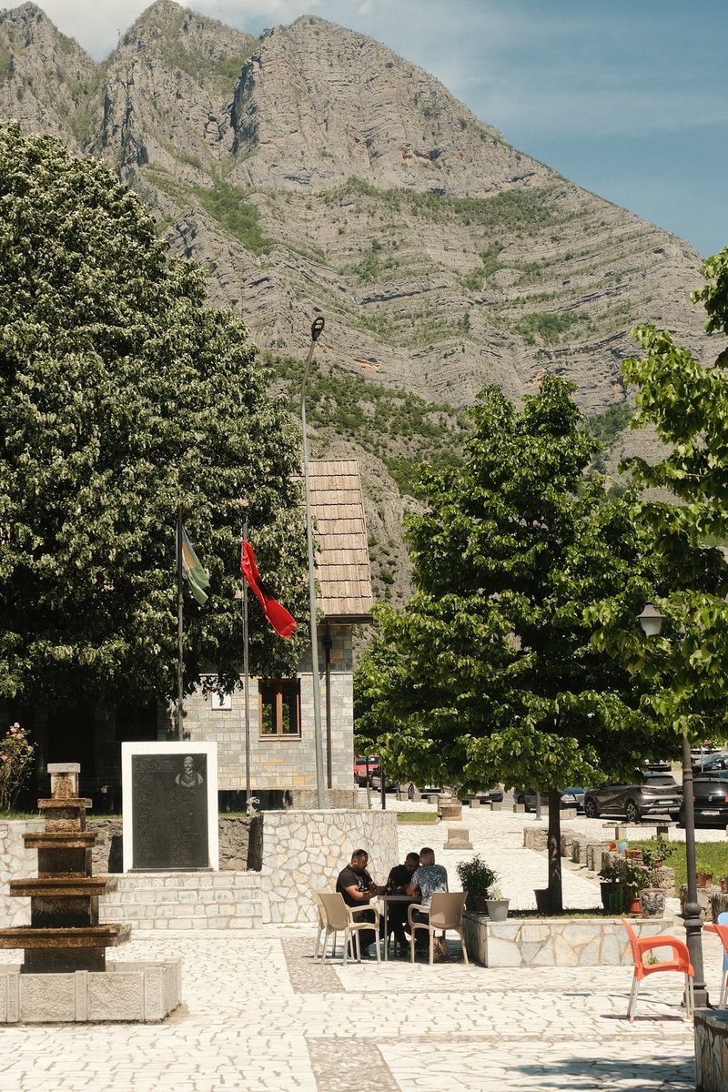 A mountainous area with a stone plaza, featuring a fountain, trees, and a building with flags and a memorial.