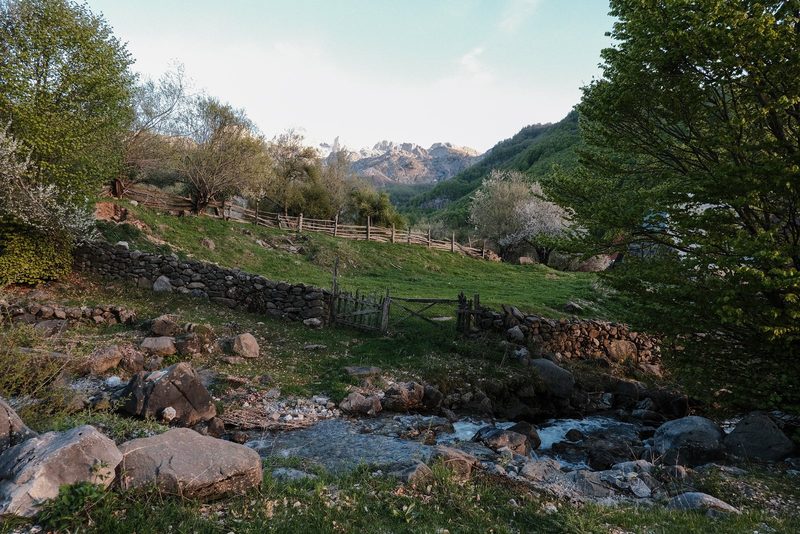 A serene landscape with a stream, rocks, and a fence in the foreground, set against a mountainous backdrop.