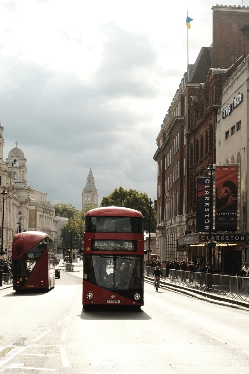 A red double-decker bus drives down a street in London, with the Houses of Parliament in the background.