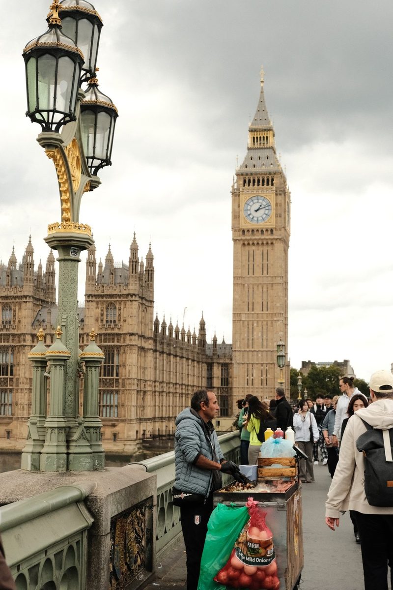 A street vendor stands on a bridge in front of the Houses of Parliament in London, with Big Ben in the background.