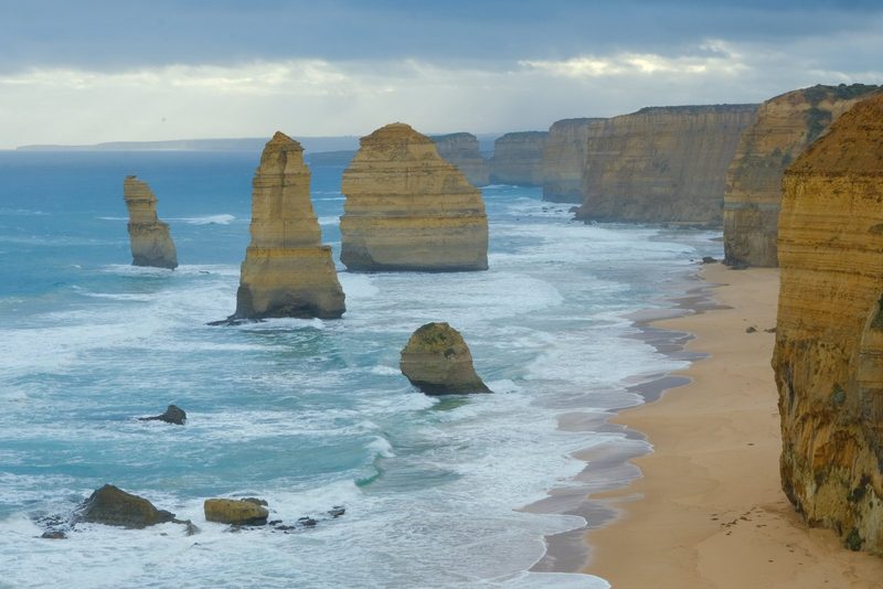 A coastal landscape featuring large rock formations in the ocean, with a sandy beach and cliffs in the foreground.