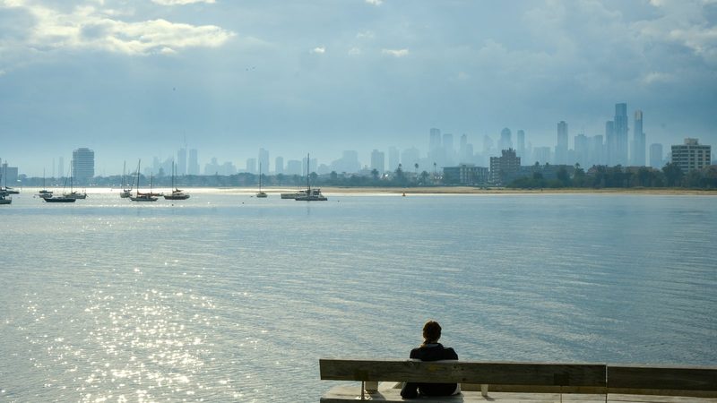 A person sits on a bench overlooking a body of water with a city skyline in the background.