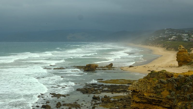 A rocky coastline with a sandy beach and a town in the background, under a cloudy sky.