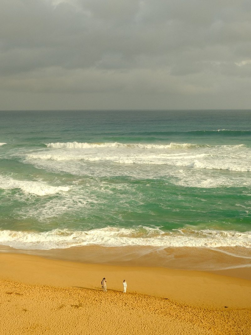 Three people in white clothing stand on a sandy beach with a large body of water and waves in the background.