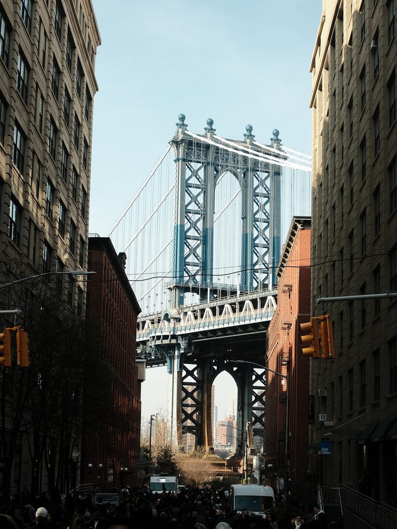 A city street with a large bridge in the background, surrounded by tall buildings and a crowd of people.