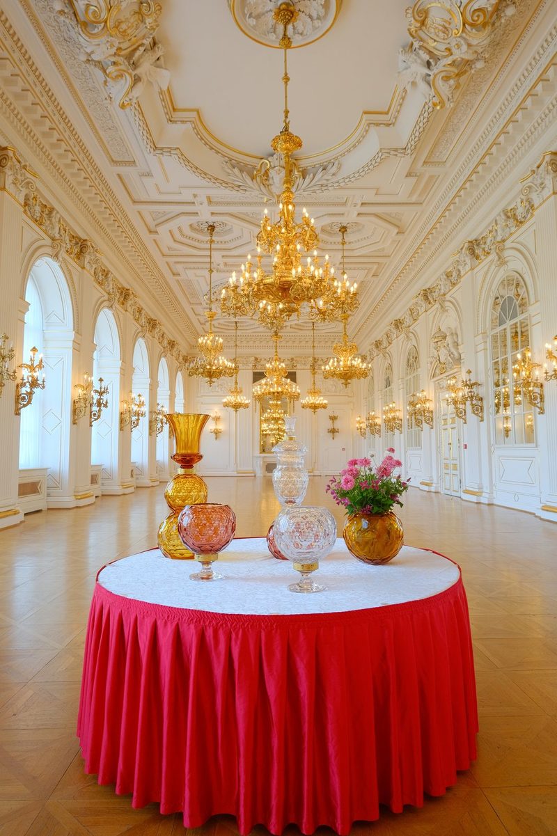 A large, ornate room with a high ceiling and chandeliers, featuring a table with glass vases and flowers on it.
