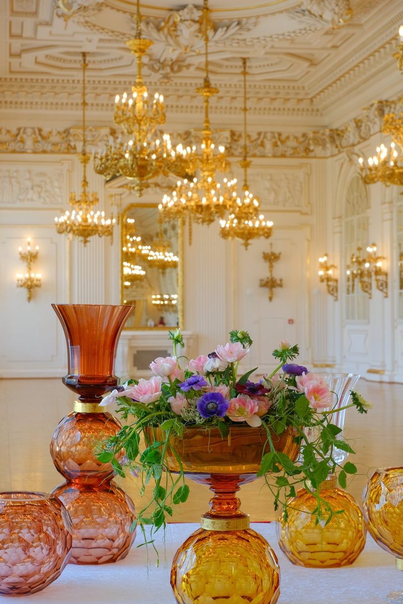 A luxurious room with ornate white and gold walls, chandeliers, and a table with amber glass vases and a floral arrangement.