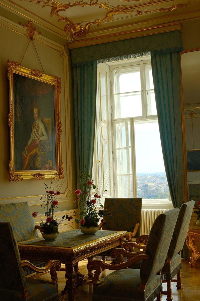 A room with a large window, a table, and chairs, featuring a portrait on the wall and a cityscape outside.