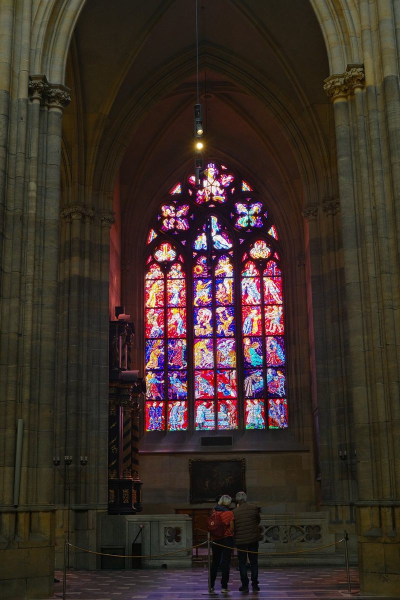 A large stained glass window in a cathedral, with people standing in front of it.
