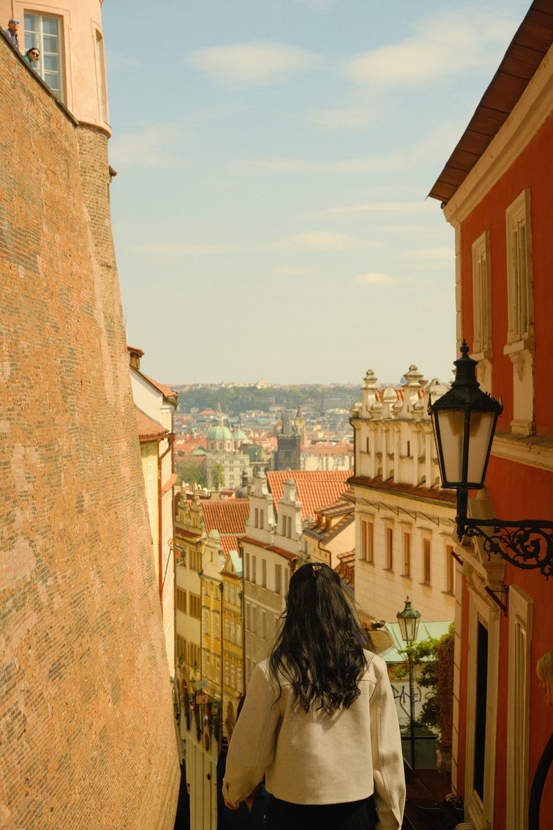 A woman with long dark hair walks down a narrow street in an old European city.
