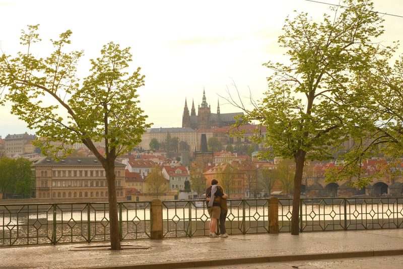 A cityscape with a river and a castle in the background, featuring people walking and standing on the riverbank.