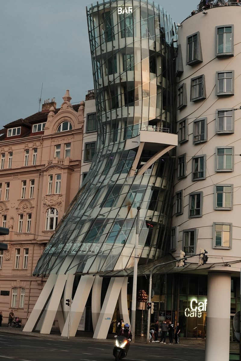 A modern glass building with a unique design stands in the middle of a city street, surrounded by other buildings and vehicles.