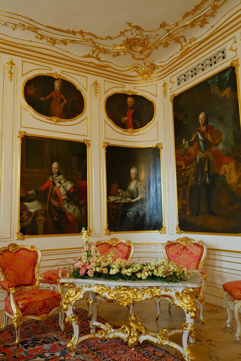A room with white walls and gold trim features five large paintings of people in formal attire, a white table with gold accents, and red chairs with gold trim.