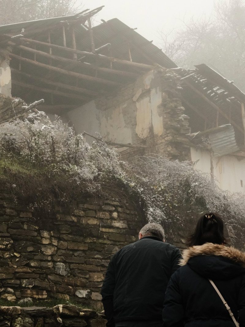 Two people, dressed in dark coats with fur-lined hoods, stand in front of a stone wall and a dilapidated building with a collapsed roof.