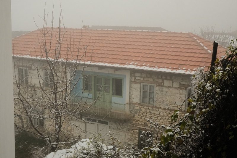 A two-story stone house with a red-tiled roof, featuring a balcony and several windows, is set amidst a snowy landscape with trees and bushes in the foreground.