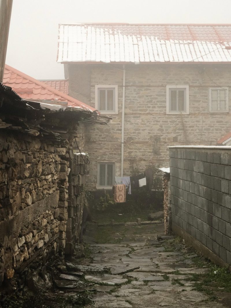 A stone-paved alleyway leads to a building with a red roof and a stone wall, featuring a clothesline with hanging items.
