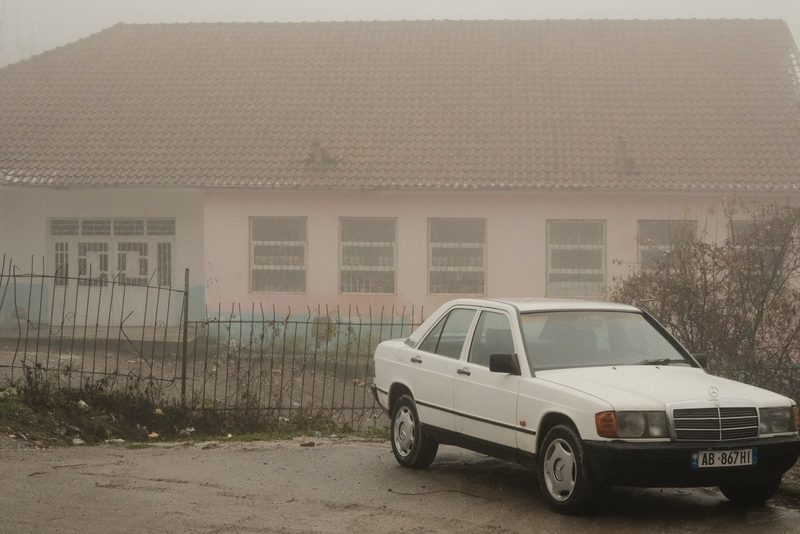 A white sedan is parked in front of a pink house with a tiled roof, surrounded by a fence and overgrown vegetation.