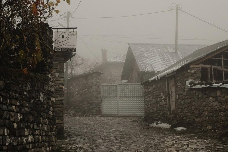 A stone wall with a sign, a gate, and a building with a metal roof, set against a backdrop of a foggy, overcast sky.