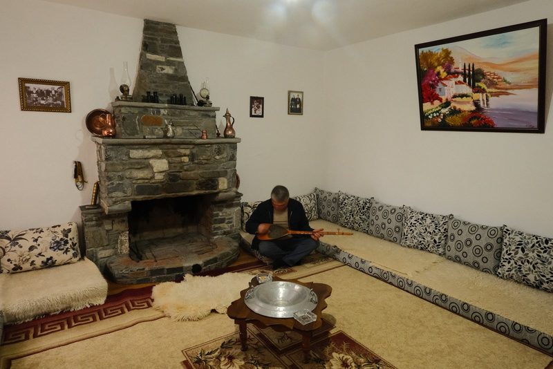 A man sits on the floor, playing a stringed instrument in a room with a fireplace and a painting on the wall.