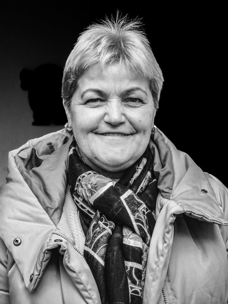 A woman with short hair and a scarf stands in front of a dark background, wearing a light-colored jacket.