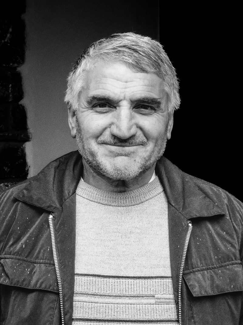 A man with short, light-colored hair and a beard, wearing a light-colored sweater and a dark jacket, stands in front of a dark background.
