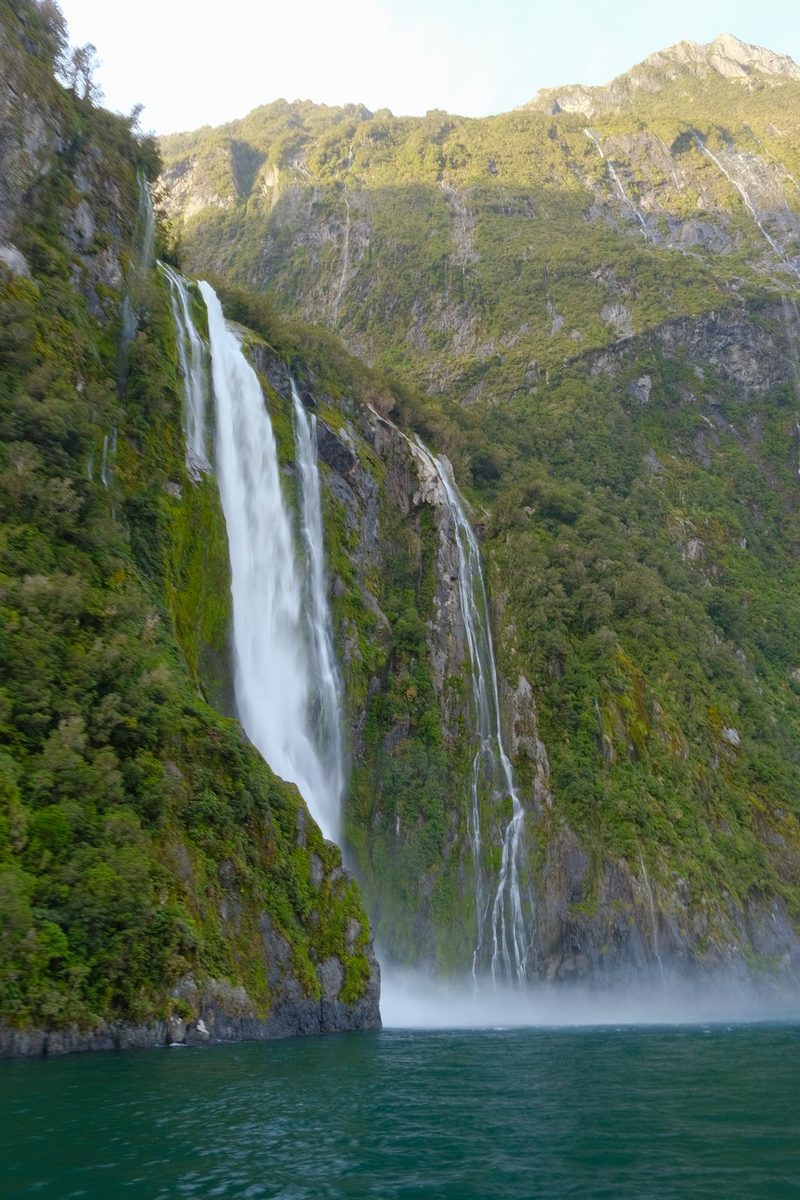 A large waterfall cascades into a body of water surrounded by lush greenery and rocky cliffs.
