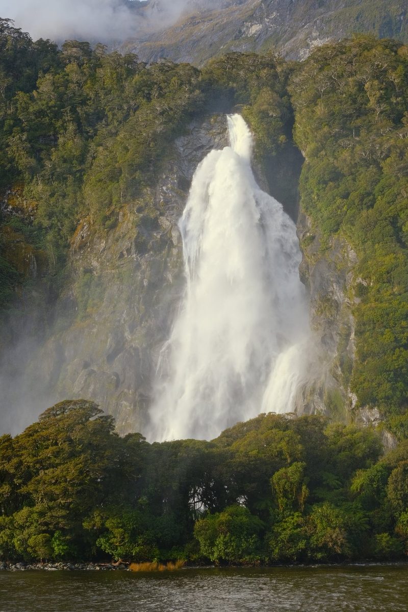 A large waterfall cascades down a rocky cliffside surrounded by lush greenery and a body of water in the foreground.