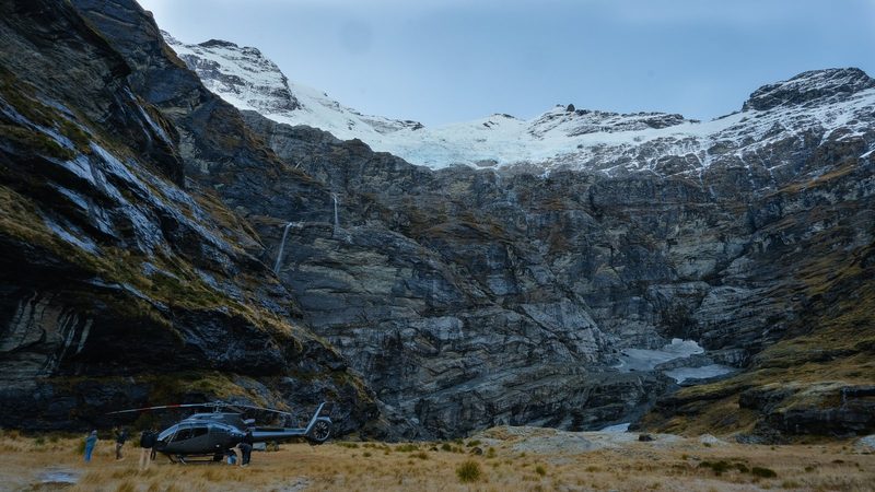 A helicopter is parked in front of a large rocky cliff face, with a group of people standing nearby, set against a backdrop of snow-capped peaks in a remote, rugged location.