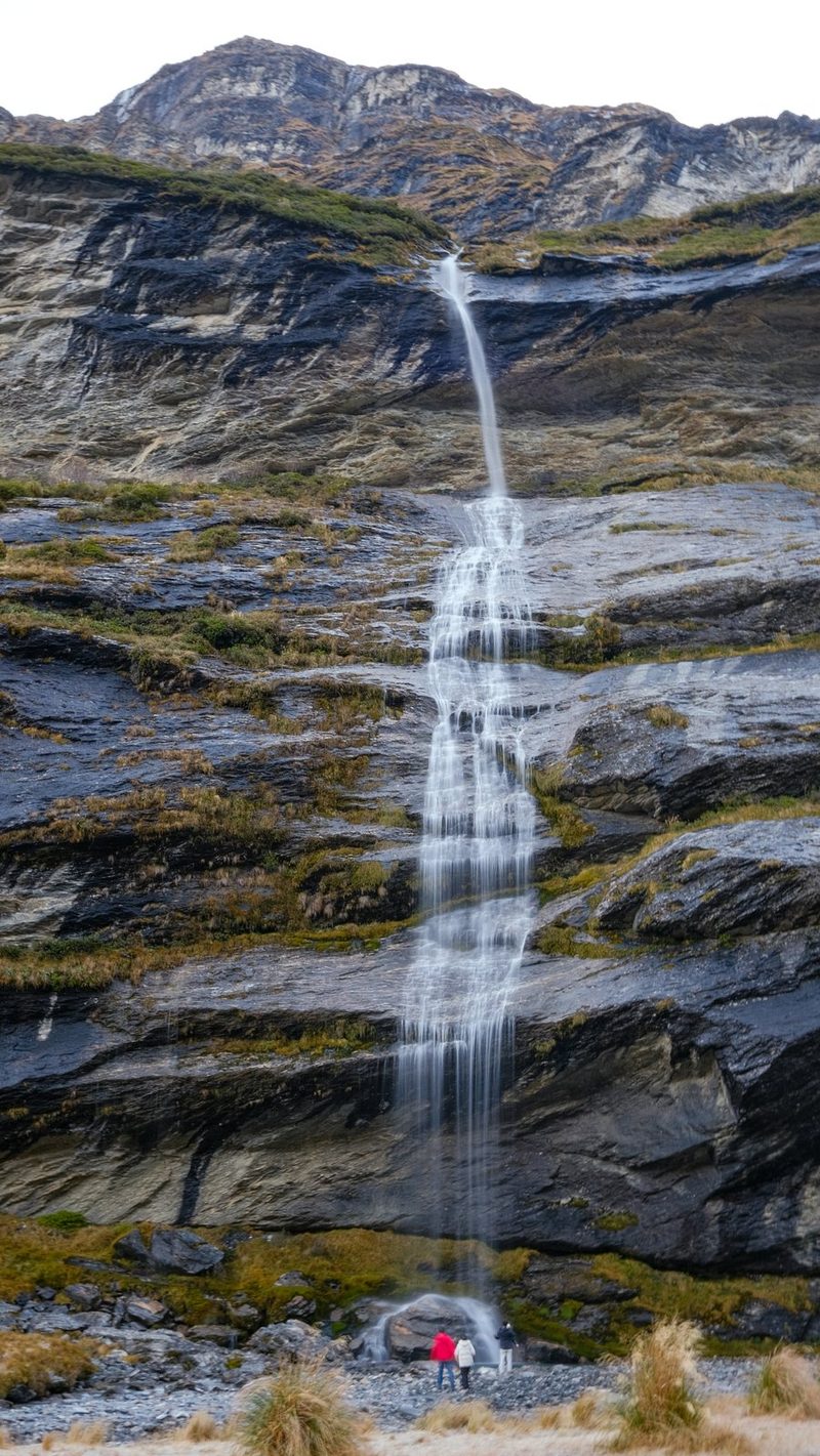 A large waterfall cascades down a rocky mountain slope, with two people standing at the base.