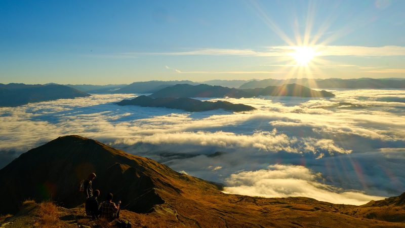 Three people sit on a mountain peak overlooking a sea of clouds and mountains in the distance.