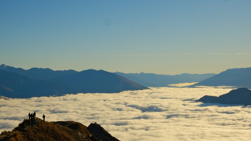 A group of people stand on a mountain peak overlooking a sea of clouds and mountains in the distance.