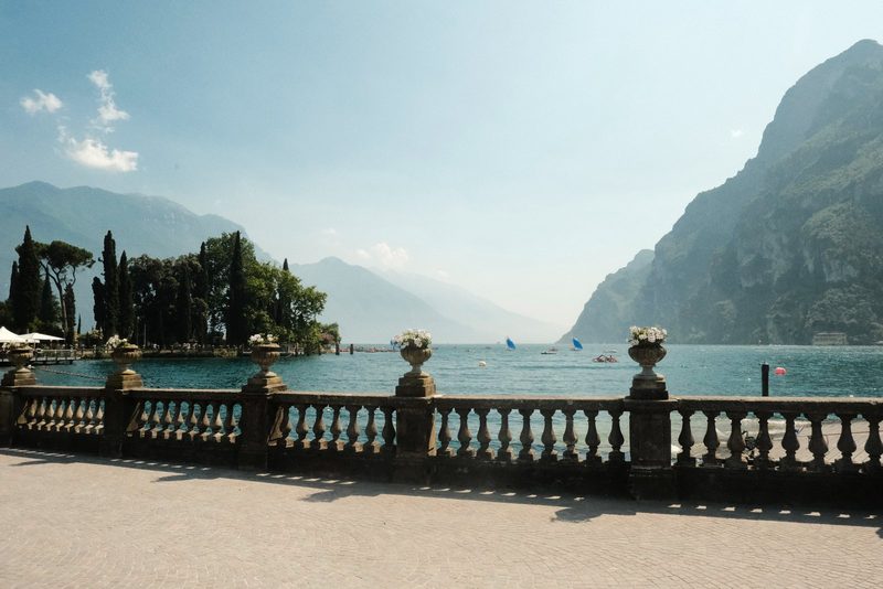A stone railing with ornate balusters and urns, overlooking a body of water with boats and a distant island.