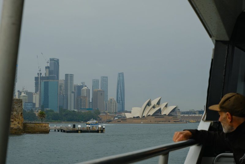 A passenger looks across the harbor toward a sail-shaped building and a dense city skyline.