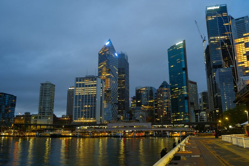 A city skyline at dusk, featuring a cluster of tall buildings with illuminated windows and a body of water in the foreground.