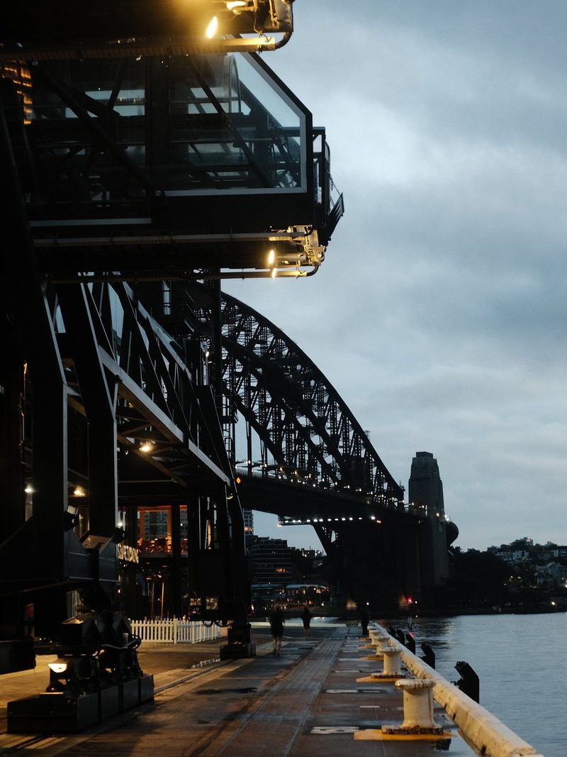 A large metal structure, possibly a bridge or crane, stands on a pier, with a walkway and railing in the foreground, and a body of water in the background.