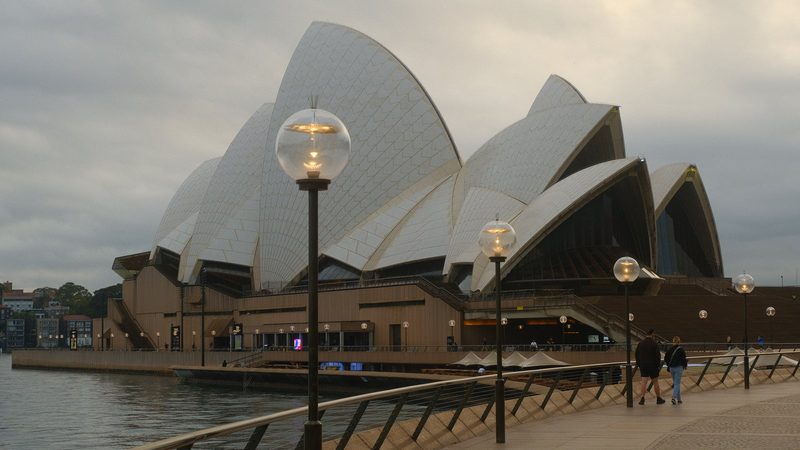 The photograph features a large building with a distinctive roof design, situated on the waterfront and surrounded by a walkway with streetlights.