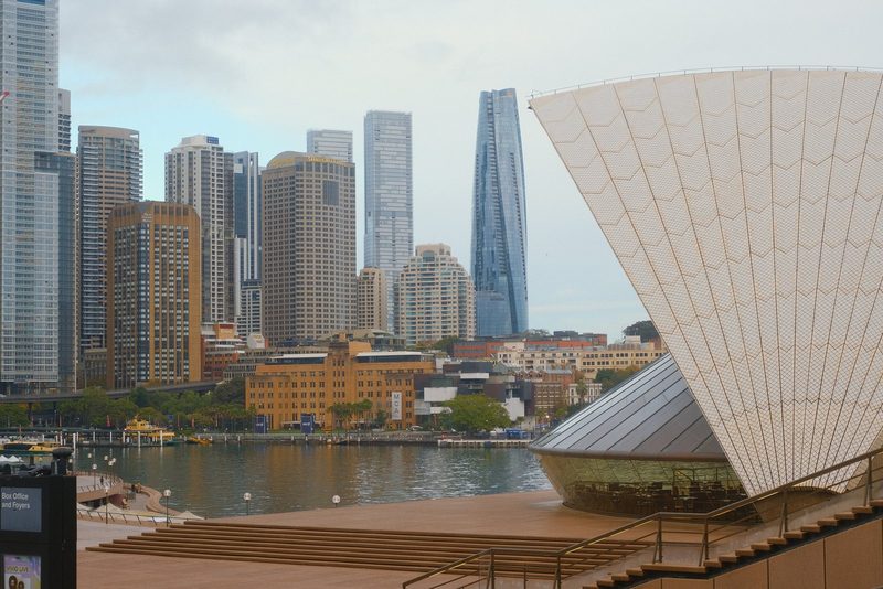 A large, curved building with a white exterior and a glass front, situated in front of a body of water and a city skyline.