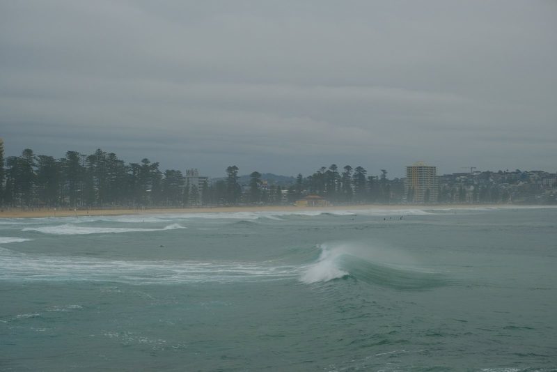 A large body of water with waves and white foam, set against a backdrop of a sandy beach and a line of trees and buildings.