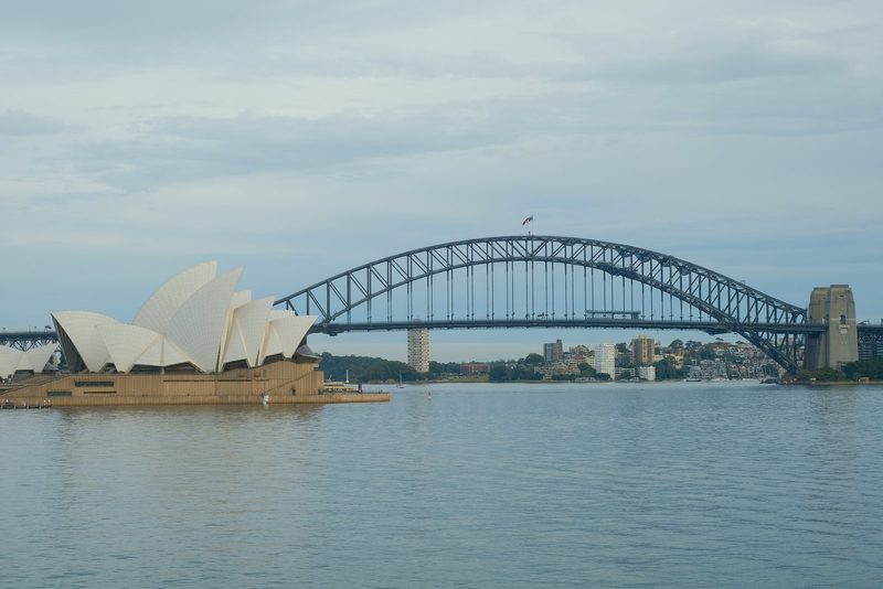 A large, arched bridge spans a body of water, with a smaller, sail-like structure on the left.