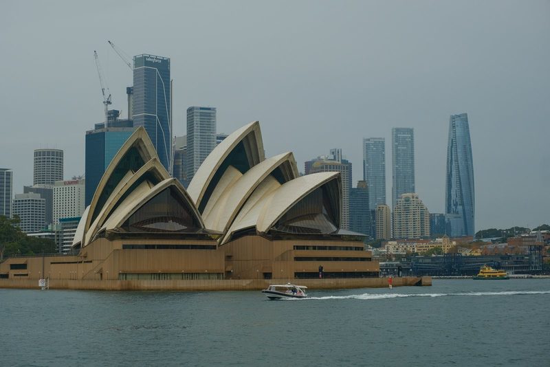 A large, modern building with a distinctive roof design, situated on the waterfront, with a boat and a ferry in the foreground.