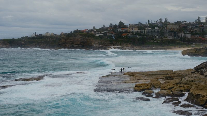 A rocky coastline with a group of surfers standing on the shore, holding surfboards, and a cliffside with buildings in the background.